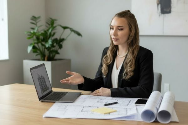 Frau sitzt am Schreibtisch mit Laptop und Bauplänen und gestikuliert während eines Gesprächs